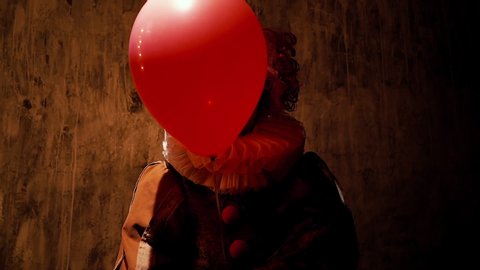 Sinister clown with colorful makeup holds balloon in his hand and smiling welcomes the viewer. Face of crazy clown is hidden behind balloon. Light shines in face. Shooting on dark red backgroundの動画素材