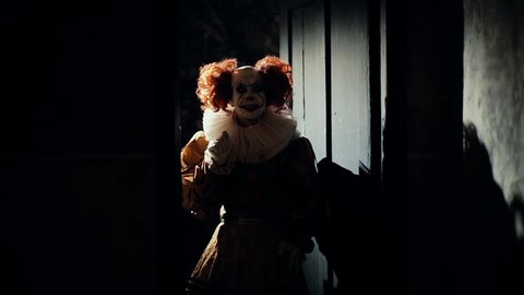 Scary clown with a crazy smile suddenly appears at the door of a dark room. Frightening clown with colorful make-up in a carnival costume waving kindly to the audience