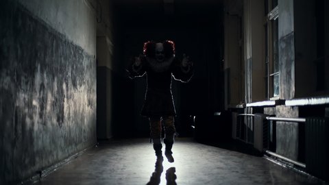 Scary clown in a dark hallway at night goes to a man in slow motion. Frightening clown with colorful makeup in a carnival costume moves his hands against the background of a bright spotlight