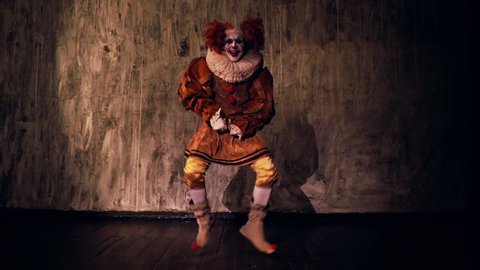 Crazy scary clown dancing against a dark red wall in darkened room. Terrible clown with colorful makeup in a festive costume. Shooting in slow motion