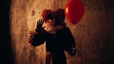 Scary clown with colorful makeup in a carnival costume, looking into the camera, holds a red balloon in his hand and welcomes the viewer in a dark room with red walls without a smile on the face.