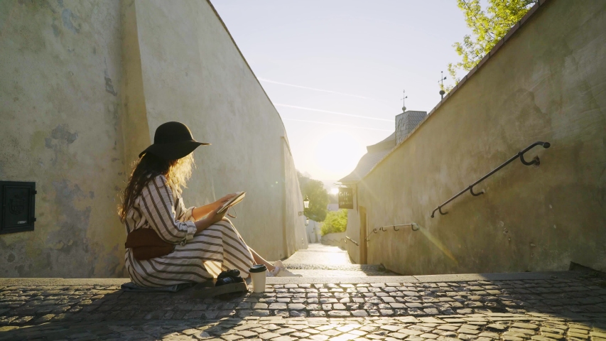 Young woman enjoying at downtown of Prague. Side view elegant lady sitting on stairway in sun shining. Lateral pan girl making notes in notepad and drinking coffee from paper mug on empty street