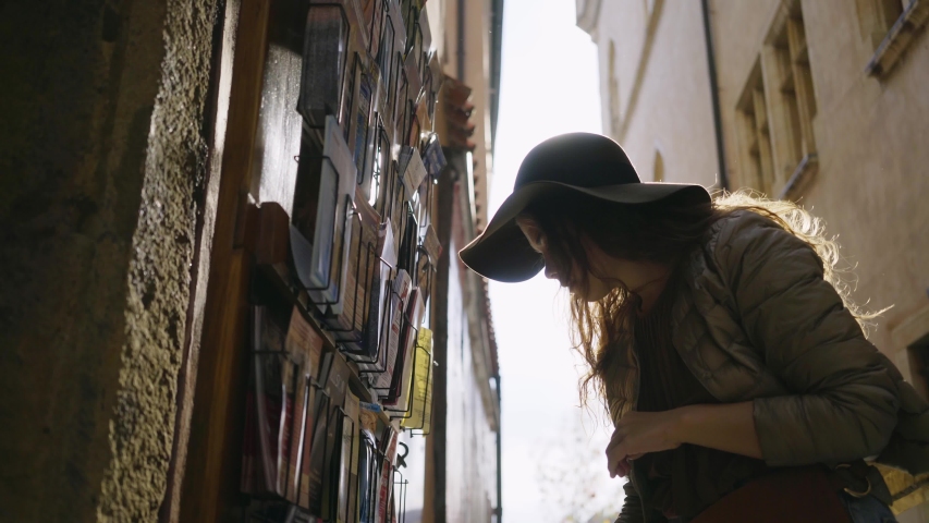 Young woman looking post cards in souvenir shop at downtown of Prague. Close-up portrait elegant girl in hat standing on street in backlit while choosing greeting cards on narrow street of old town