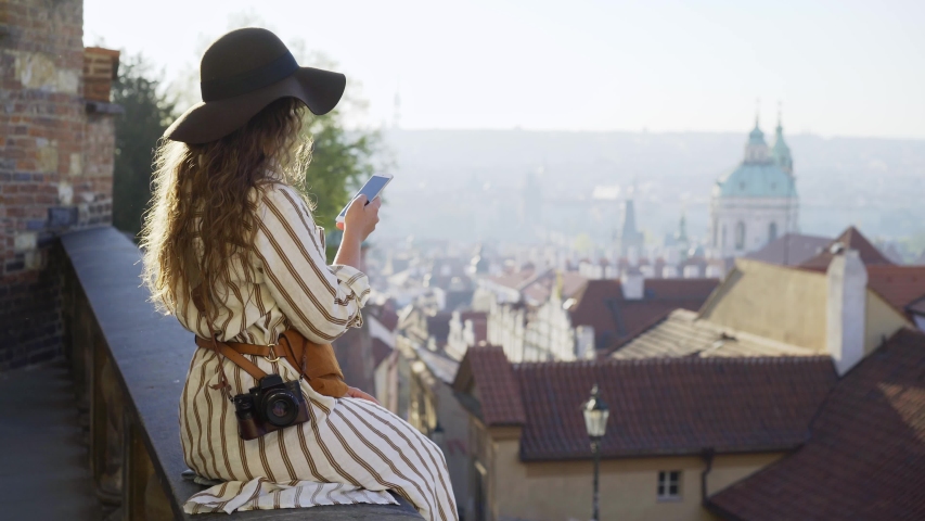 Side view of casual woman sitting on view point above downtown of Prague.Young girl tourist enjoying beautiful view red tiled roof, belfry tower and cathedrals on top of hill at sunny day