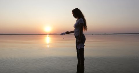 Silhouette of a girl with long curly hair letting a few paper boats go on the water. Toy ship sailing to the red sunset. Young woman dreaming during sunset. Vídeo Stock