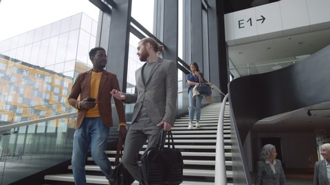 Low angle view of handsome African man wearing casual clothes and Caucasian young man with red hair and beard going together downstairs in airport while chatting: stockvideo