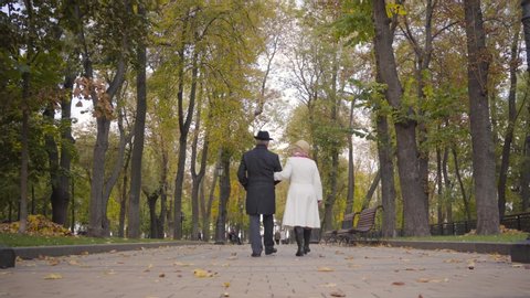 Back view of a mature Caucasian couple walking in the autumn park and talking. Mature man in black clothes and his wife in white coat strolling along the alley.