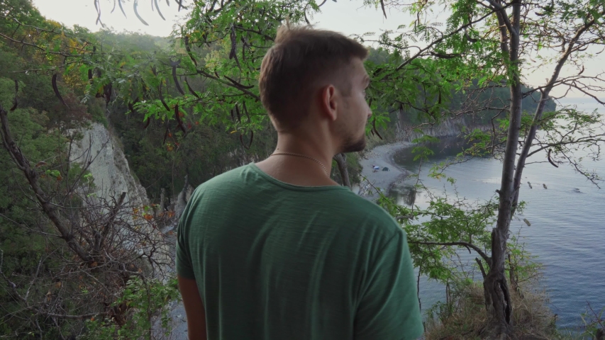A young man from behind goes to the cliff and looks at the bottom.