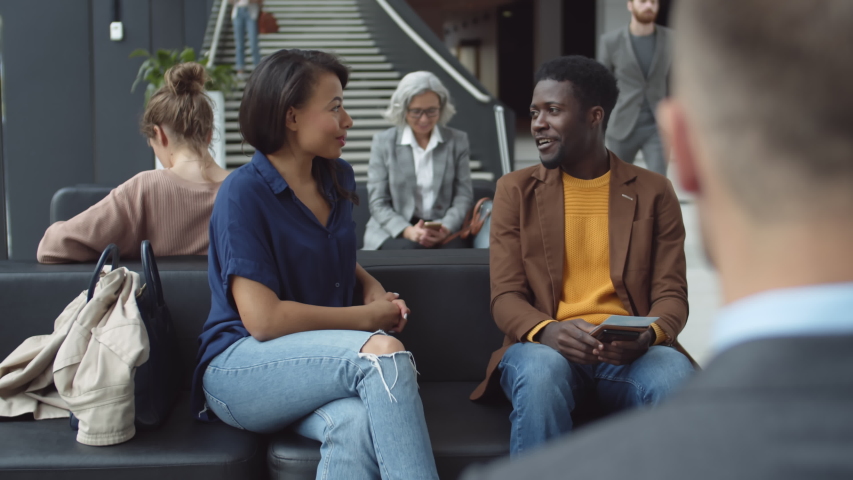 Medium shot of African man wearing casual clothes sitting in lobby with tickets in his hands and chatting with mixed-race pretty woman sitting nearby | Shutterstock HD Video #1038757673