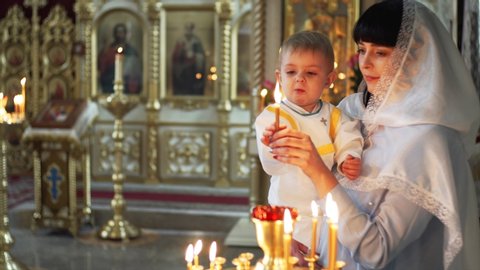Portrait of mother and her cute con with burning candle in cathedral, Russia. Icons in golden frames are on the background