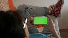 Girl Using Touchscreen Mobile Phone. Young Woman Home Lying on a Couch with Green Screen Smartphone in Horizontal Mode. Girl Using Smartphone, Browsing Internet, Watching Video Content, Blogs. POV.