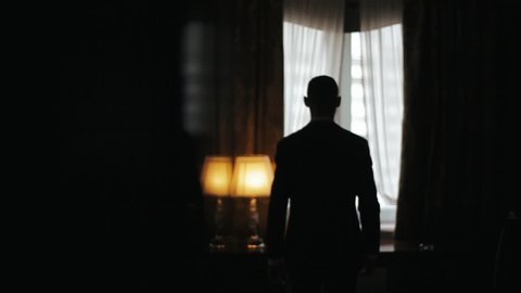 Silhouette of a Man wearing suit walking from a dark room towards a bright window in slow motion. Man stops, puts his hands on a table and looks pensive to the window.