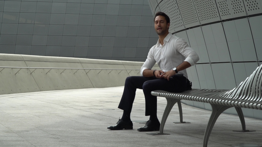 Wide shot of handsome and fit European man with dark hair in white collared shirt and dark trousers is sitting in an impressive modern architectural setting putting his phone away