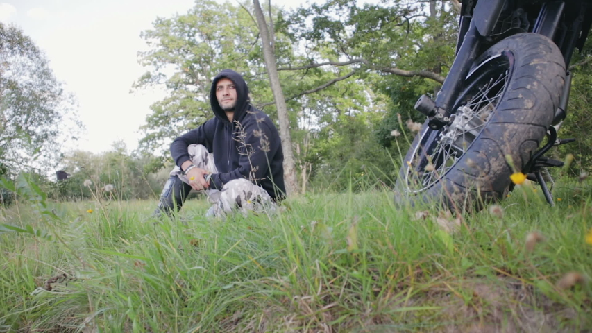 A handsome guy in a black hood, in protective boots and khaki pants sits and breathes fresh air in the forest near his motorcycle. Adventure tour, Enduro and off-road, summer day. 