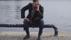 Young man in overcoat using a mobile phone outdoors while sitting on a waterfront promenade on a metal bollard