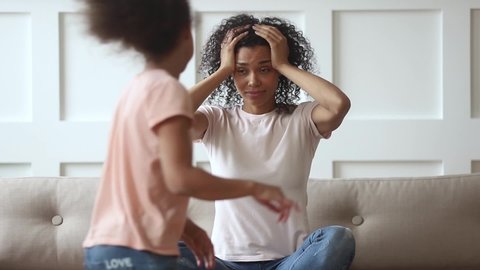 Noisy girl and boy play at home while nanny sit on sofa, african son and daughter running around tired mom under stress holds head suffers from restless children, need calm tiredness from kids concept