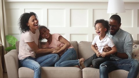 African mixed-race cheerful full family sit on couch in living room play with little kids active loving parents tickling children daughter and son funny spending time together at home activity concept
