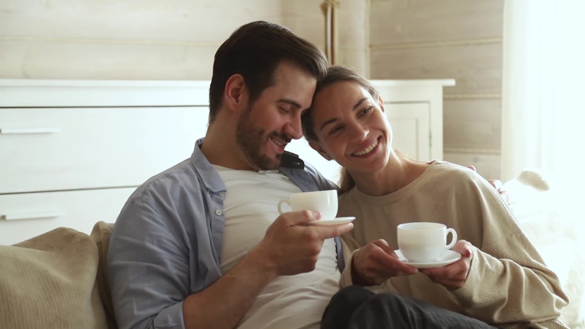Lovely newlyweds married couple in love sit on couch in living room talking feels happy, wife comfortably settled on the husband lap holding tea cups drink morning coffee enjoy cozy warm conversation