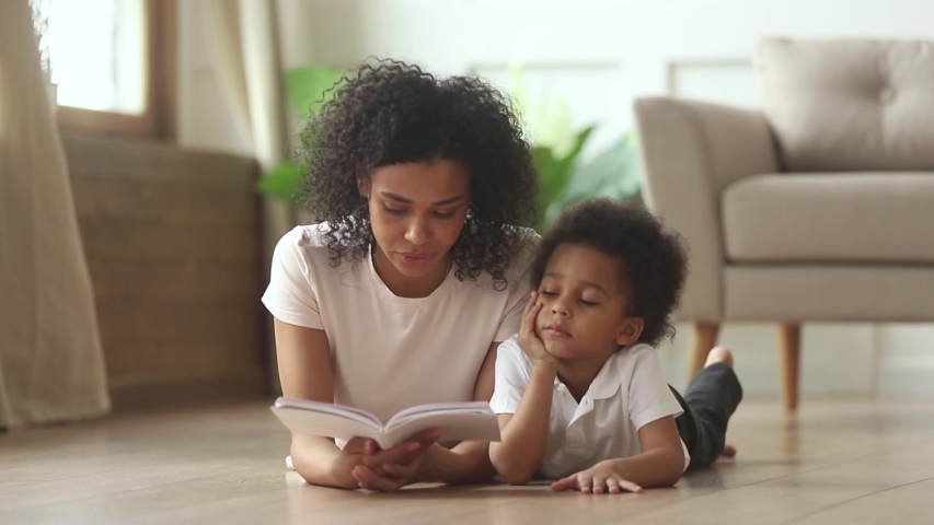 Mixed-race toddler little son lying on warm floor at modern home in living room listens black mother hold book reading fairy tale story feels bored disinterested, free time, development of kid concept | Shutterstock HD Video #1038796346