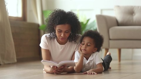 Mixed-race toddler little son lying on warm floor at modern home in living room listens black mother hold book reading fairy tale story feels bored disinterested, free time, development of kid concept