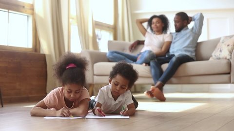 Little mixed race african siblings lying on warm wooden floor in living room drawing with colorful pencils while young black mother and father resting, talking on couch activity at modern home concept