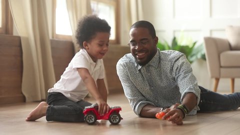 Caring african dad and little son sitting lying on warm wooden floor with underfloor heating play with toy cars spend enjoy time together at home, modern comfy house, leisure activity with kid concept
