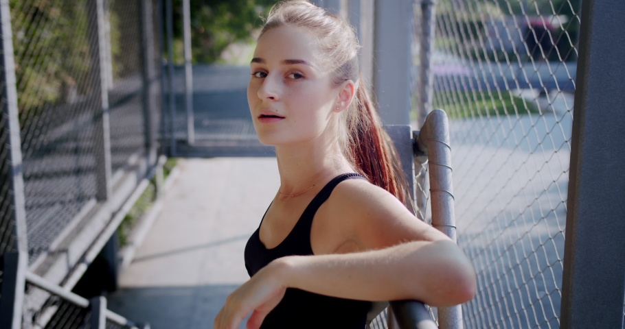 Close up of cute healthy young female leaning on metal fence and smiling looking to camera, leisure summer morning of happy nice girl resting outdoor after power workout and physical activity