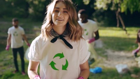 Attractive female volunteer picking up trash waste in the park with ecology team. Portrait of nice-looking smiling girl helping environment cleaning forest nature with friends.