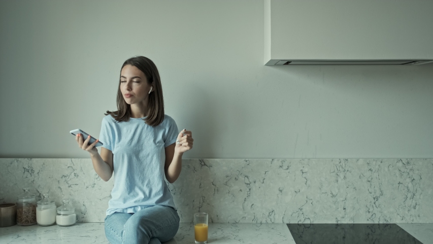 Attractive happy young brunette woman listening music with wireless earphones on smartphone and singing while sitting at the kitchen table at home