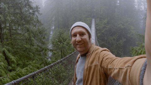 Young man hiking in green forest takes selfies on suspended bridge 
