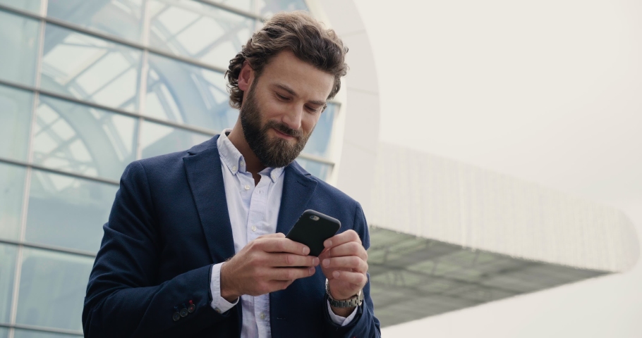 Handsome Businessman browsing his Smartphone near modern Office Building. Attractive Man is smiling and wearing Smart-casual Style. Social Networking. Luxury Lifestyle. Smartphones. Apps.