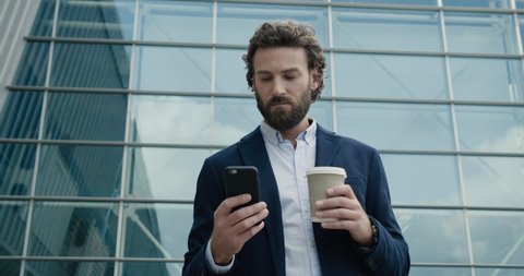 Serious Businessman is texting Messages on his Smartphone near Office Building. Stylish Man is drinking coffee to go and browsing his Phone while the Break. Office Man. Startup Life. Social Network.