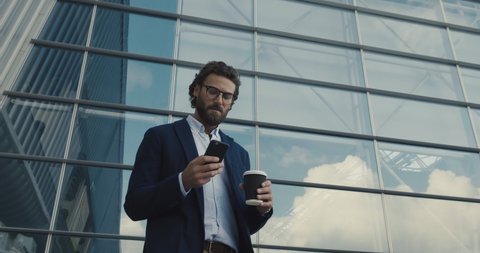 Young Multinational Man Chatting in Glasses working on his Smartphone. Typing a Message. Walking by the Big Industrial Building. Drinking coffee to go. Smiling Sincerly. Classical Jacket. Apps.