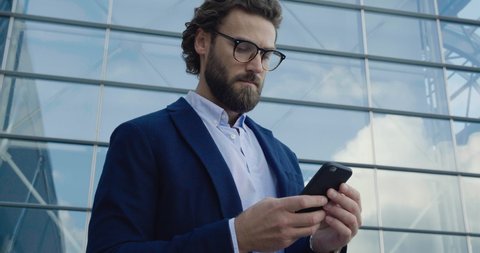 Young confident Professor in Glasses looking to the Camera confidently. Portrait of young Caucasian Man with stylish hair and Look. Using his Smartphone. Social Network. Work outside. Success. People.