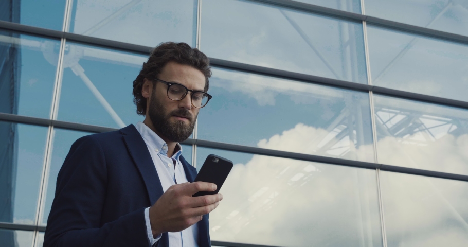 Handsome bearded Startup Businessman in Glasses using mobile Phone. Young man texting Messages, cheating on his Smartphone while walking in Office District. Business. Social Networking. People. Apps. | Shutterstock HD Video #1038809891