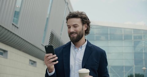 Happy bearded Businessman is texting Messages on his Smartphone and smiles. Young Man is walking near modern Office Building while drinking Coffee to go. Business. Social communication. People.