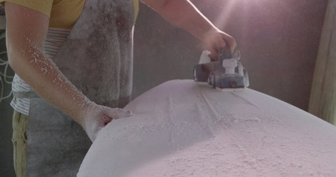 Side view mid section of a millennial Caucasian male surfboard maker in his studio, sanding a new surfboard with a tool during the manufacturing process