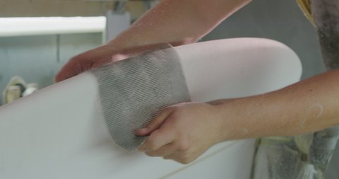 Close up of the hands of a millennial Caucasian male surfboard maker in his studio, rubbing the edges of a surfboard with a mesh sanding gauze during the manufacturing process