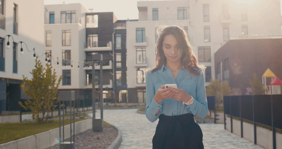 Camera zooming in on the young pretty Caucasian girl who standing in sunlight in the living area in the town among buildings and tapping on the smartphone. Outdoors. | Shutterstock HD Video #1038821891