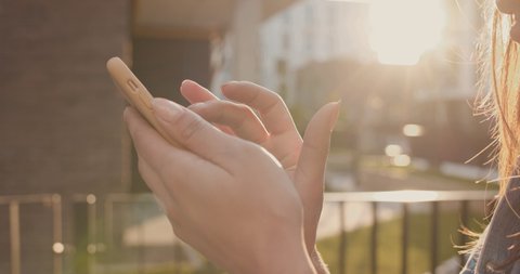 Close up of the smartphone device in hands of the young Caucasian woman in sunlight while she tapping and texting on is screen. Outdoor.