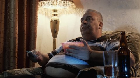 Fat lazy elderly man eating chips and drinking beer while watching TV