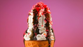 Ice cream cone close-up. Colorful sprinkles falling on ice cream with chocolate syrup topping. Icecream in waffle cone, rotating on pink background. White Sweet dessert closeup, rotation. 4K UHD
