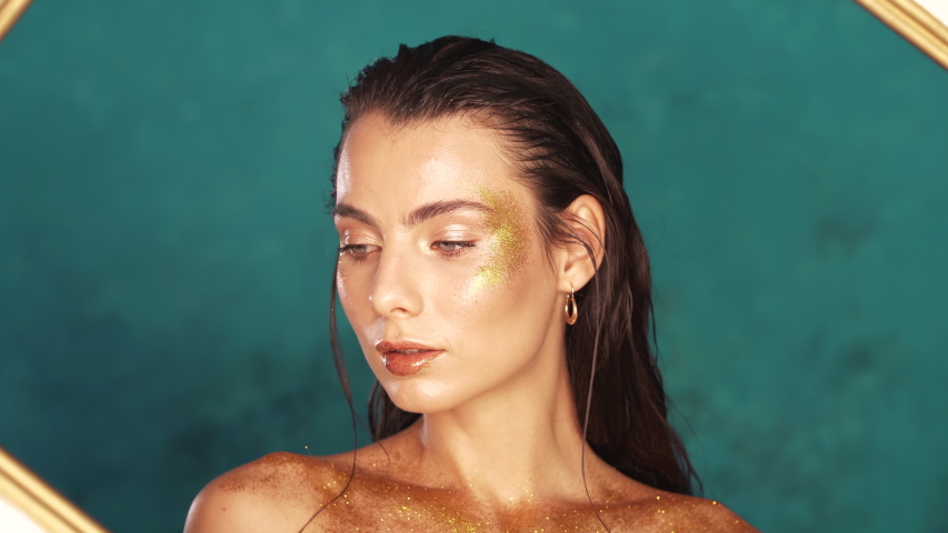 Amazing fashion model girl close-up portrait with picture frame in turquoise studio. Golden makeup, visage, body art concept.