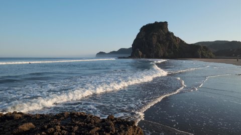 Piha, Auckland Region, New Zealand - April 20, 2019: Morning surf at South Piha beach with Lion Rock in the background. Two people, one taking a walk the other surfing.