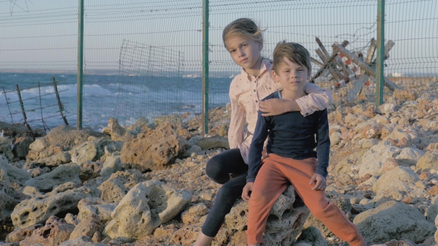 two poor suffering orphan refugees children boy and girl sitting on stone near sea with state border with migration razor barbed fence behind