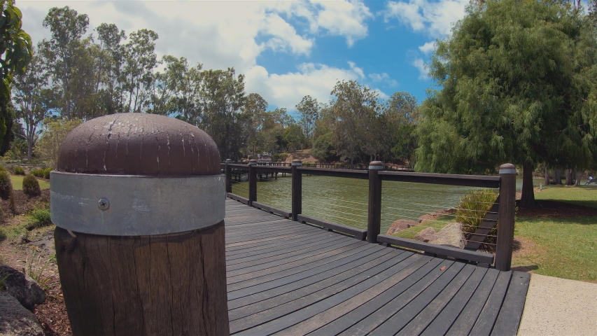 Wooden Bridge Over Lake. Beautiful Scenic Outdoor Timber Walkway & Curved Board Walk Over Calm Tranquil River Water. Lake Bridge View & Peaceful Waterfront Landscape. Grass Lawn Trees & Sunny Blue Sky