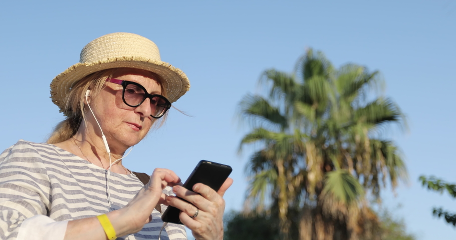 Blonde elderly woman in sunglasses and straw hat choosing a song on mobile phone. Portrait of sixty years old woman enjoying music in earphones. Golden hour with palm and blue sky on the background.