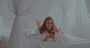 Nice girl lies on a bed in a cozy bedroom and communicates by video call with her friend. Young happy woman speaks on the smartphone in the room.