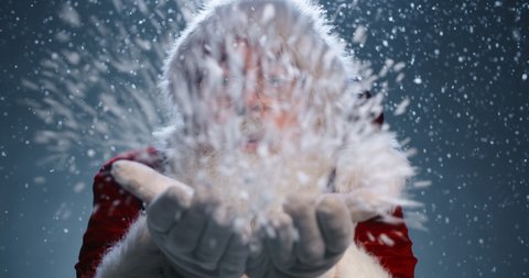 Bearded old man in Santa Claus clothing smiling and blowing snow into camera, isolated over blue background - christmas spirit concept close up 4k footage