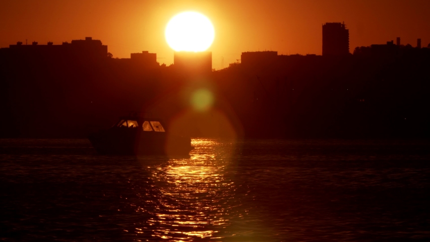 Bright shot across the bay: silhouette of motor boat, cityscape of Vladivostok, yellow sky because of rising sun. Summer sunrise. Russia